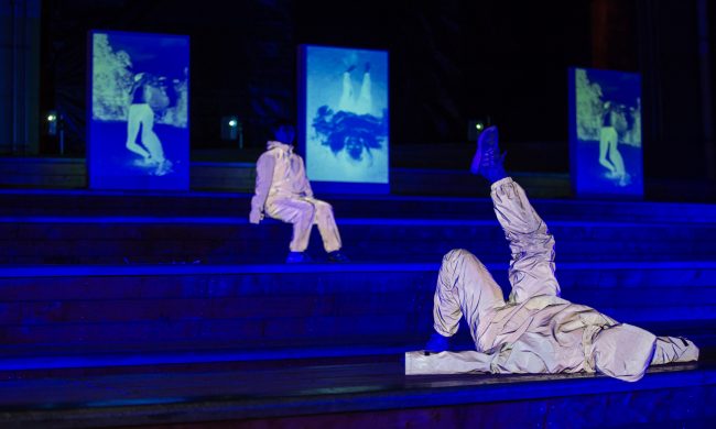 Photo in blue light of two performers sitting and laying on stairs wearing reflective suits.