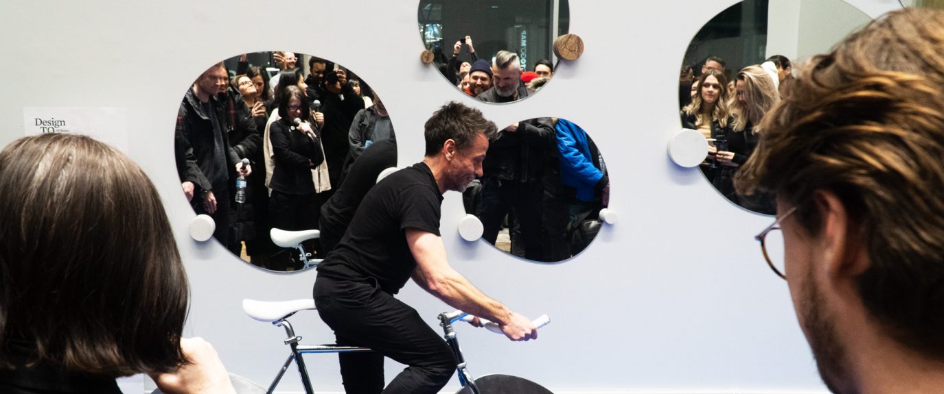 Photo of a man in a black t-shirt and black pants riding a stationary bike in front of a group of onlookers.