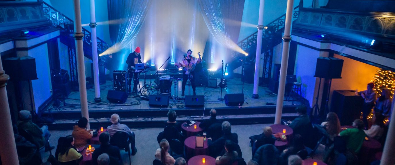 Photo of an indoor event with 2 musicians performing on a stage and audience seated at tables.