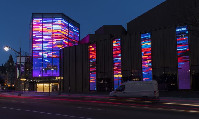 Photo of Canada’s National Art Centre taken from the street at night with lights on all windows on display lit up in full colour.