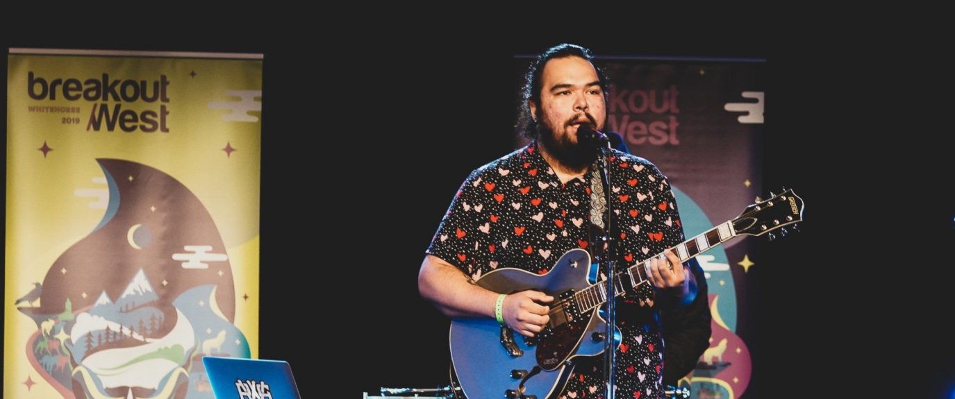 Musician with a blue guitar and a microphone in front of them. The have dark black hair that is pulled back and a beard. They are wearing a button up shirt with polka dots and pink and red hearts. There is table behind them with a laptop and a turntable. Two BreakOut West pop up banners are behind the table.