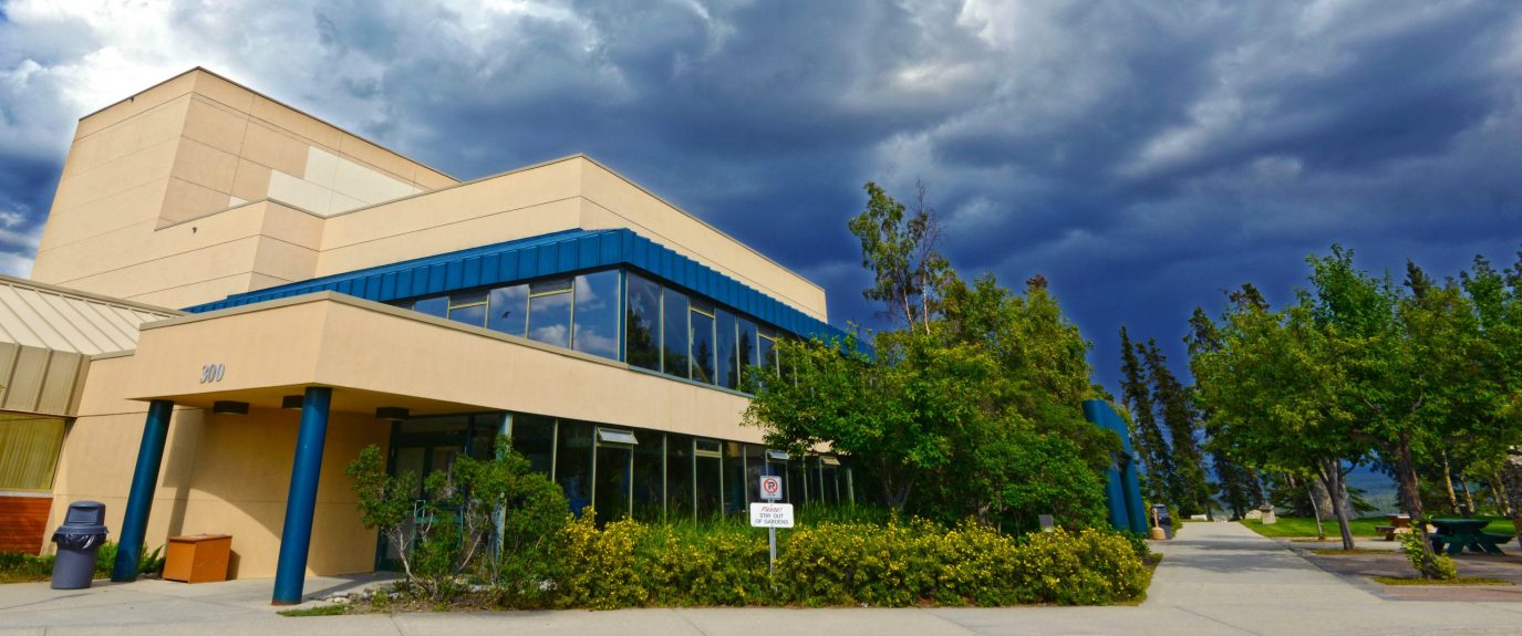 Exterior of the Yukon Arts Centre, a building made of beige concrete, blue pillars and detailing with lots of windows surrounded by green trees and bushes. The sky in the background is cloudy and dark blue.