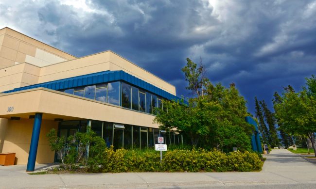 The outside of the Yukon Arts Centre building. The building is a beige and blue colour with lots of windows. It is sounded by green trees and bushes. The sky is cloudy and a dark blue.