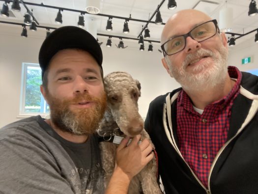 Two bearded men standing indoors with a short-haired beige dog being held by the man on the left. The room is white and has a lighting grid on the ceiling.