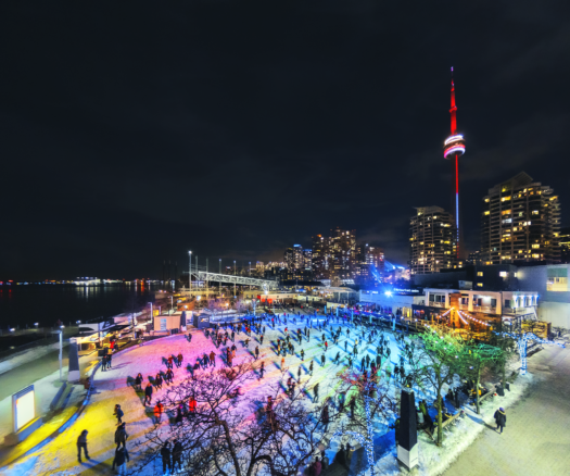 People skating on outdoor rink lit with multi-coloured lights at night. Harbourfront Centre’s main building and Toronto’s skyline with the CN Tower is in the background.