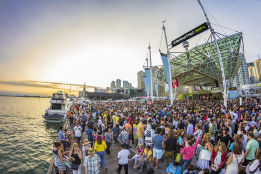 Open-air concert stage beside Lake Ontario at dusk with a large crowd gathered. A band is performing on the stage which features a large Harbourfront Centre sign.