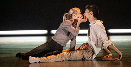 Two dancers sitting on a stage in grey and beige clothing. The dancer on the left uses one hand to hold the face of the other dancer, leaning towards them. The backdrop is black with bright white lights along the floor.