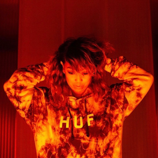 A sepia toned half-shot of artist Cell7 wearing a tie-dye pattern hooded sweater with the word HUF on it in white. The artist's arms are behind their head tilting it downwards.