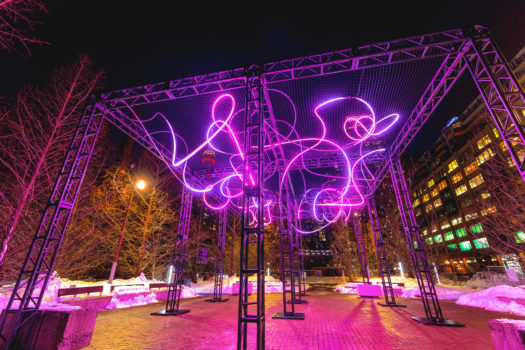 Outdoor light art installation at night. The strings of light are thin, purple and swirly, supported by tall metal truss columns and wire mesh.