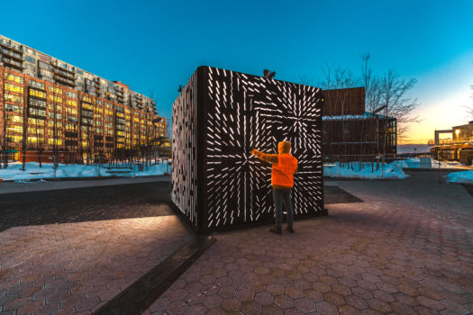 A person in an orange jacket and grey pants standing in front of a large cube light installation at dusk. The cube has thin white lights with the person reaching their hand towards it and appearing to make lines of light radiate in all directions from where their hand is placed.