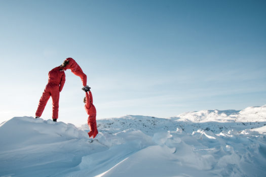 Three circus performers in red long-sleeved jumpsuits form an upside-down U shape atop a snowy mountain range in daytime.