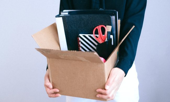 Person in a black long sleeve shirt and white pants holding a cardboard box filled with miscellaneous office supplies.
