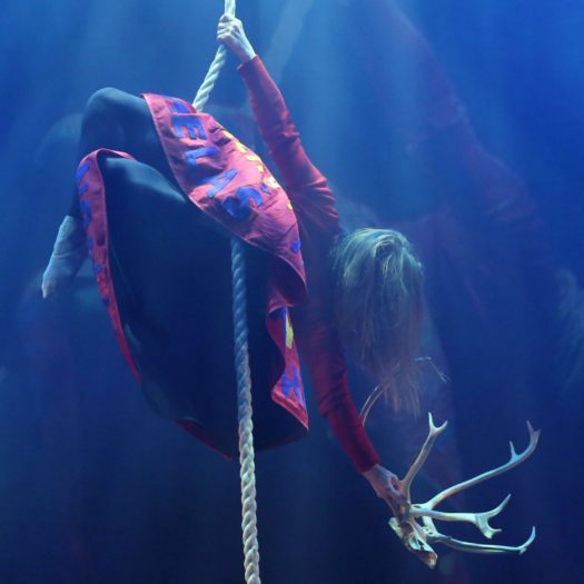 Aerialist Camille Therese Karlsen wearing a red long-sleeve shirt and skirt hangs upside down with her legs and one arm holding onto a thick rope. Her free hand holds a reindeer skull.