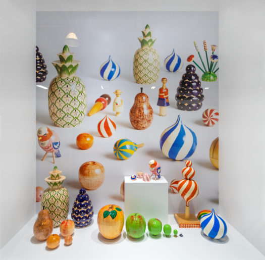 A white vitrine features brightly coloured painted and lacquered wood carvings in the shapes of fruit, including pineapples, pear, apple and apricot. Next to the fruit is a white cube with a pink lacquered bird perched on top.