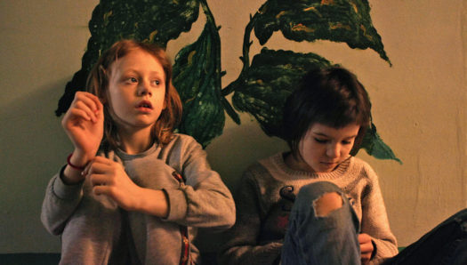 Two children with shoulder length hair wearing grey sweaters sit against a beige wall with a leaf-like mural painted on it.