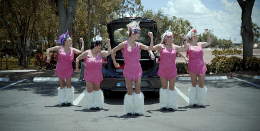 Five women in their 60's wearing knee-high white fur boots and pink fringe dresses pose in a v-formation in front of a car with an open trunk in a parking lot with trees in the background.