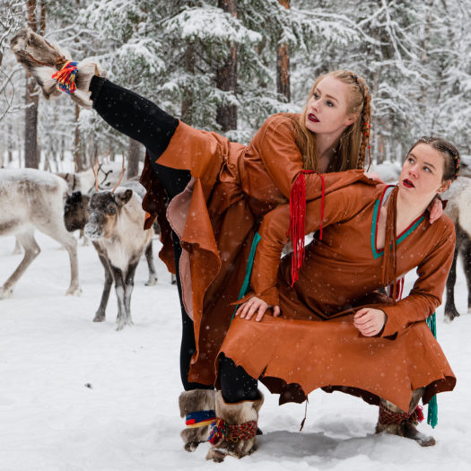 Two dancers with braided hair wearing brown leather long-sleeve dresses, fur boots and black pants in a snowy evergreen forest with reindeer behind them. The dancer on the left has a raised leg and leans on the other dancer for support.