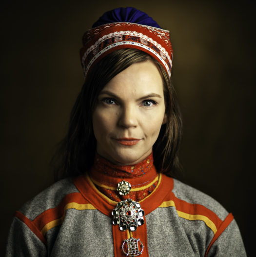 Headshot of artist Sara Marielle Gaup Beaska, a woman with brown hair and blue eyes wearing grey, yellow and red gákti, traditional Sámi clothing.
