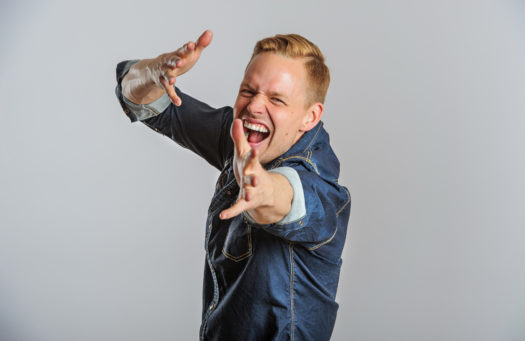 Upper body shot of rap artist Signmark with his arms stretched out towards the camera and a open-mouthed animated facial expression. He has short blonde hair and blue eyes and wears a dark denim long-sleeve button-up shirt.