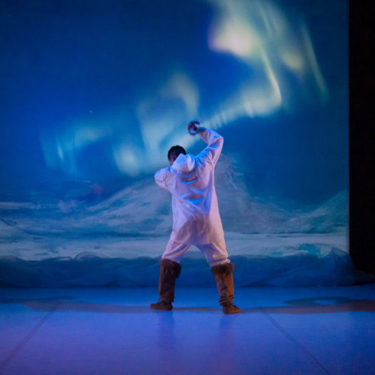 A performer stands facing upstage with one arm raised and legs slightly apart wearing a white long-sleeve jumpsuit with a hood and knee-high brown leather boots. The stage backdrop shows a snowy mountain range with blue aurora borealis lights at night.