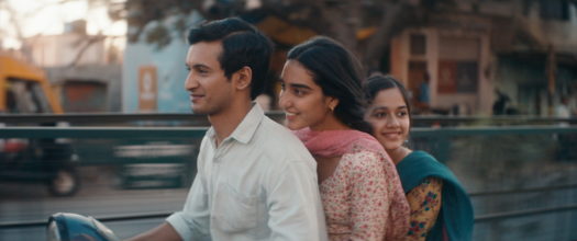 Three people on a motorcycle with a blurred background. The driver wears a white collared button-up shirt and the passengers wear colourful saris.