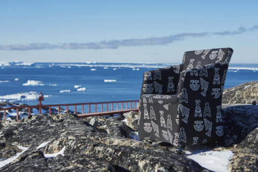 An armchair placed on a rocky, snowy shore near a body of water in daytime. The chair is black with white Inuk-inspired designs.