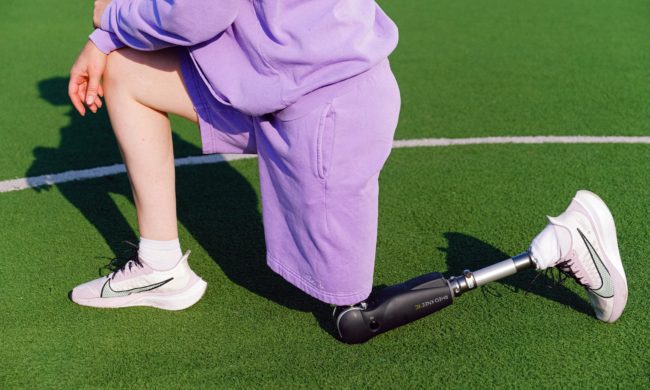 Close-cropped image of a person in a purple tracksuit with a leg prosthesis kneeling on a green AstroTurf sports field.