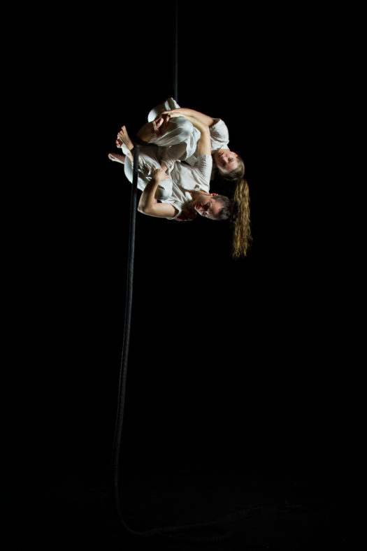 Two performers with long hair tied into high ponytails wear white short-sleeve shirts and pants while hanging from a rope sideways with their limbs wrapped around the rope.