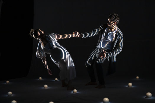 Two performers in loose white costumes lean away from each other and counterbalance by holding the opposite hand of the other. The stage is dark with white striped lighting on the performers and small white spheres placed around them on the floor.