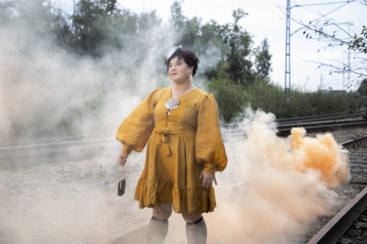 Artist Sara Ajnnak stands on an outdoor railway track wearing a mustard yellow knee-length dress with orange smoke surrounding her.