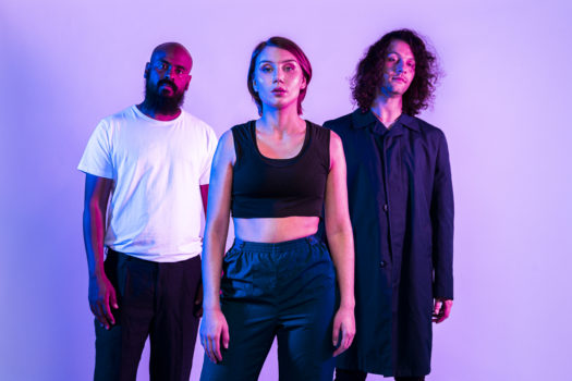 Members of music trio ISÁK stand facing the camera in a triangle formation all wearing neutral coloured clothing with purple tinted-lighting against a purple backdrop.