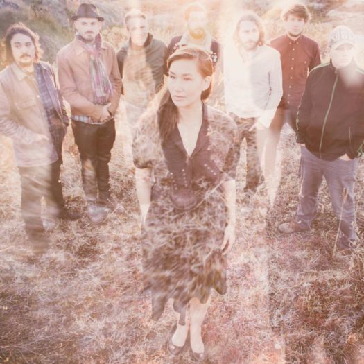 A sepia tone image of singer Nive standing in a grass-covered field wearing a dark short-sleeve collared dress with seven members of her band standing in a semi-circle behind her.