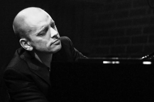 Black and white image of pianist Tord Gustavsen sitting behind a piano with his eyes closed, feeling the music, in a black suit jacket.