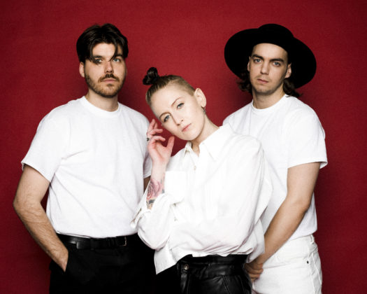 Upper body shot of music trio Vök standing in front of a red backdrop, each wearing a white shirt and black pants, with one member wearing white pants and a black round brim hat.