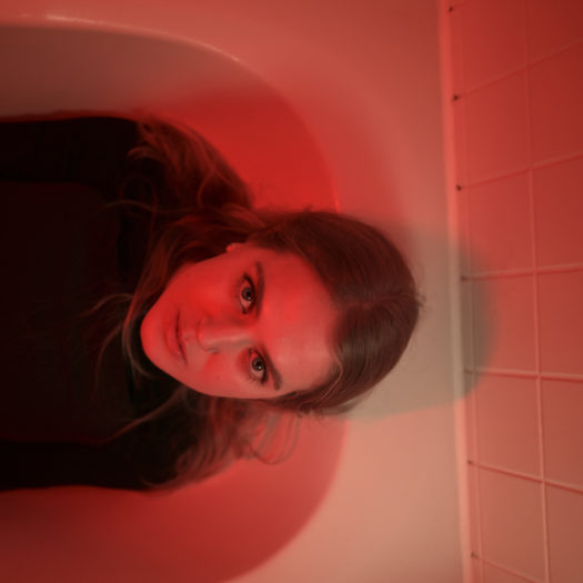 Artist leoblu, a woman with middle-parted shoulder length hair and blue eyes wearing black eye liner and a black top sitting in a white bathtub with red lighting. The image is turned sideways.