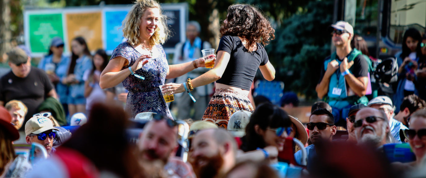 Two folkies are pictured getting their groove on amongst the crowd at the Calgary Folk Music Festival. They are both dancing and holding beverages in Big Rock cups.