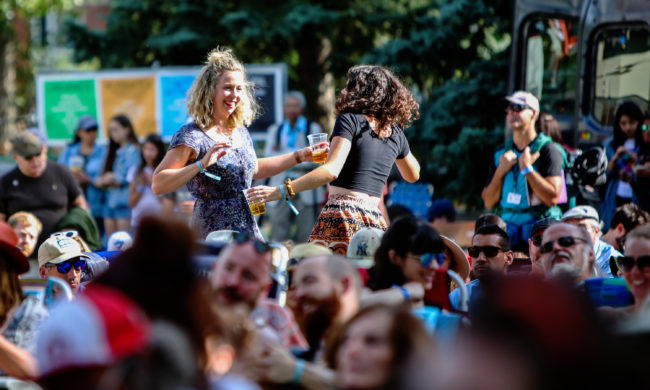Two folkies are pictured getting their groove on amongst the crowd at the Calgary Folk Music Festival. They are both dancing and holding beverages in Big Rock cups.