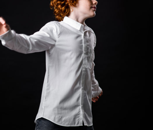 A young dancer with curly red hair wearing a white dress shirt twirls with his arms open as if to accept a hug.