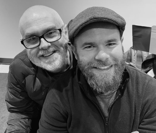 Black and white image of two bearded men smiling widely, with one slightly behind the other in an indoor space.
