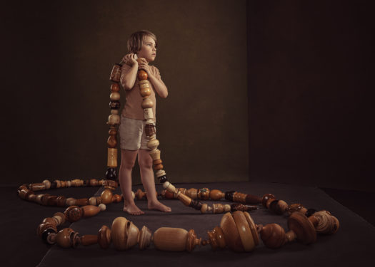 A barefoot child in neutral coloured shorts and shirt holds a chain of large wooden beads that coil around on the floor against a brown background.