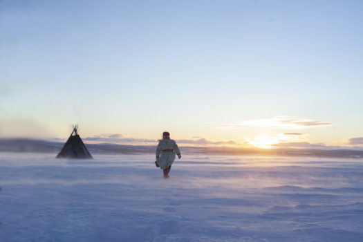 A person dressed in traditional Sámi fur attire walking on a snowy flatland towards a setting sun and a tent called a Lavvu.