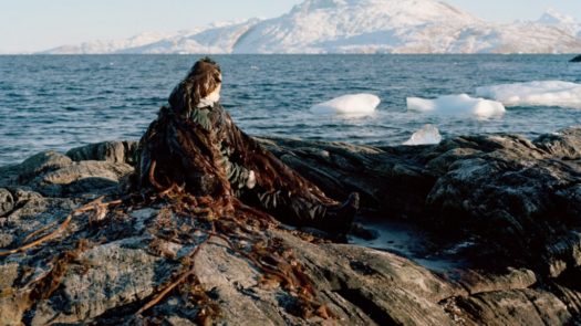 A person sits covered in seaweed on a rocky shore next to a body of water with pieces of ice floating in it and a mountain in the background in daytime.