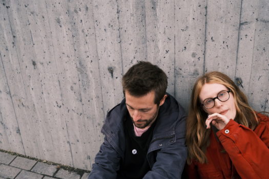 Music duo BSÍ pictured from the waist up, sitting against a concrete wall. Silla Thorarensen is on the right wearing round black glasses and a burnt orange coat. Julius Pollux Rothlaender is on the left wearing a grey coat.