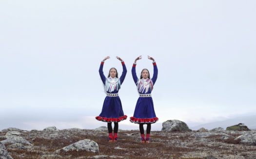 Two dancers stand on a flat rocky and grassy surface both wearing traditional Sámi attire, gákti, with their arms raised above their heads to create an oval shape.