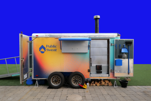 The Mobile Sweat sauna, a large four wheel trailer with the words Public Sweat written on its side set-up outdoors on cobblestone against a blue background.