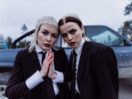 Half-shot of two women Rebecka and Sanna, members of The Magnettes trio. Both wear oversized black suits with a black tie and white collared shirt, and have mid-length blonde hair with dark makeup.