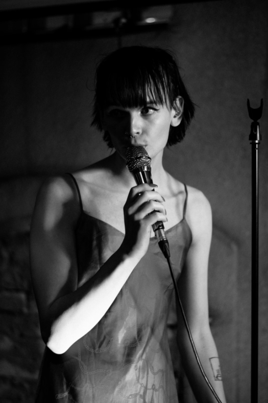 Black and white image of artist Kid Vicious with dark chin length hair and bangs wearing a spagetti strap dress and singing into a microphone.