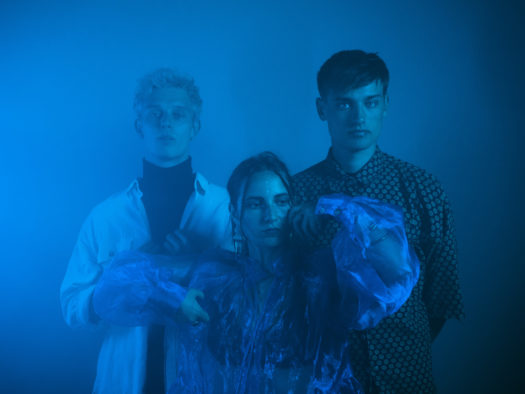 AySay music trio stand facing the camera in various styles of long-sleeve shirts with blue lighting and a blue backdrop.