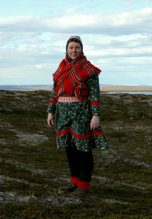 Music artist Ánnámáret stands outdoors on a rocky, grassy plain on a partially cloudy day wearing red, green and black traditional Sámi attire, gákti.