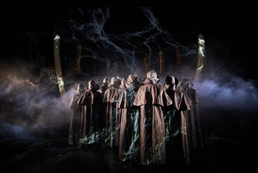 A group of performers stand in a V formation wearing black cloaks and hats, facing upstage towards whale bone props on a dark, hazy stage.